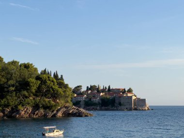 Sveti Stefan adasına giden küçük bir tekne yeşil ağaçlarla kaplı kayalık bir tepenin etrafından geçiyor. Karadağ. Yüksek kalite fotoğraf