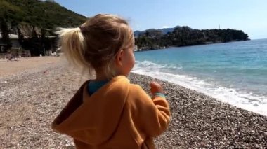 Küçük kız sahilde duruyor ve deniz sörfüne bakıyor. Yüksek kalite 4k görüntü