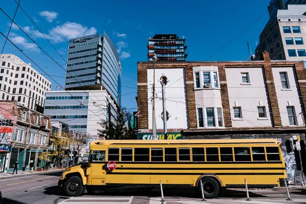 Ön planda sarı bir okul otobüsünün yer aldığı canlı bir şehir caddesi, eski tuğla yapılarla açık mavi gökyüzü altında modern yüksek binalar arasında çelişiyor. Yüksek kalite fotoğraf