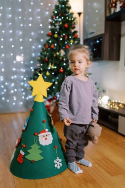 Küçük kız oyuncak bir Noel ağacının yanında oyuncak bir ayıyla duruyor ve başka tarafa bakıyor. Yüksek kalite fotoğraf