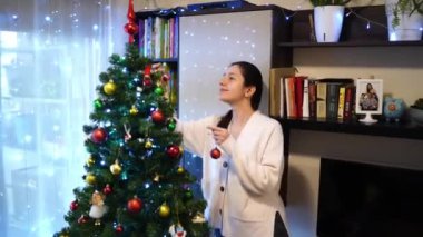 Genç kadın odadaki Noel ağacına parlak toplar asıyor. Yüksek kalite 4k görüntü