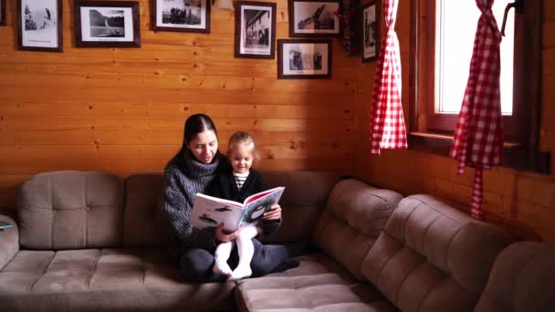 ママは木製の家に大きなソファーに座っている小さな女の子に本を読みます 高品質の4K映像 — ストック動画