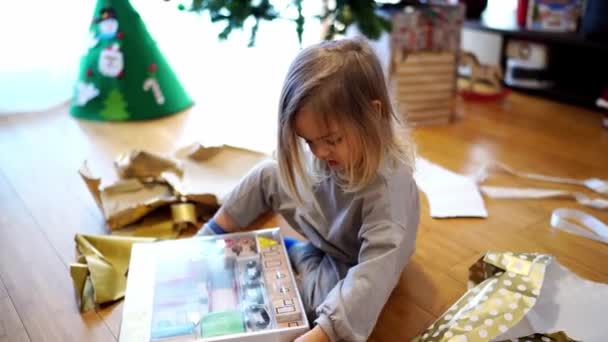 小さな女の子はクリスマスツリーの近くに座っている間 贈り物から光沢のある包装紙を取り除きます 高品質の4K映像 — ストック動画