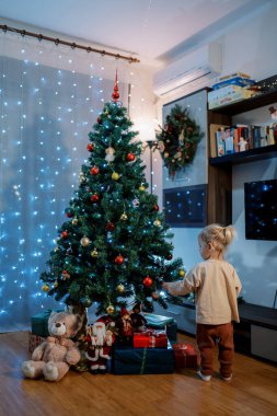 Küçük kız süslü bir Noel ağacının yanında duruyor ve toplara dokunuyor. Arkadan bak. Yüksek kalite fotoğraf