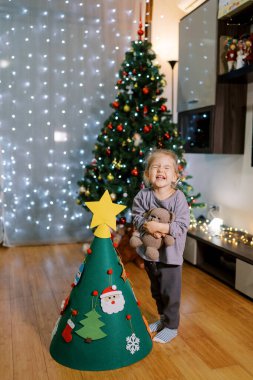 Oyuncak bir Noel ağacının yanında oyuncak ayıyla dikilen küçük kız gözlerini kırar ve yüzünü buruşturur. Yüksek kalite fotoğraf