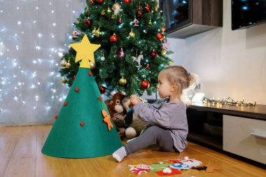 Küçük kız, odanın zemininde otururken oyuncak bir Noel ağacına keçeli bir oyuncak asıyor. Yüksek kalite fotoğraf