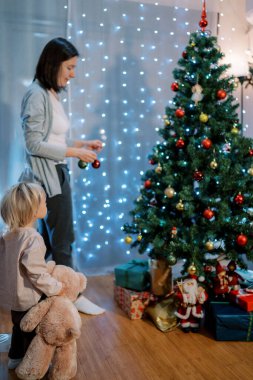 Elinde oyuncak ayıyla küçük bir kız annesinin Noel ağacını süslemesini izliyor. Arkadan bak. Yüksek kalite fotoğraf