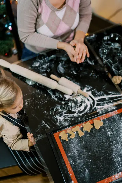 Küçük kız masada annesinin yanına oturur ve bir tepsi kesilmiş kurabiyeye bakar. Üst Manzara. Yüksek kalite fotoğraf