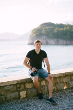 Sırt çantalı fotoğrafçı deniz kenarındaki taş bir çitin üzerinde oturuyor. Yüksek kalite fotoğraf