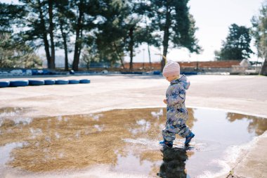 Lastik çizmeli ve tulumlu küçük bir kız kaldırımda büyük bir su birikintisinde yürüyor. Yan görüş. Yüksek kalite fotoğraf