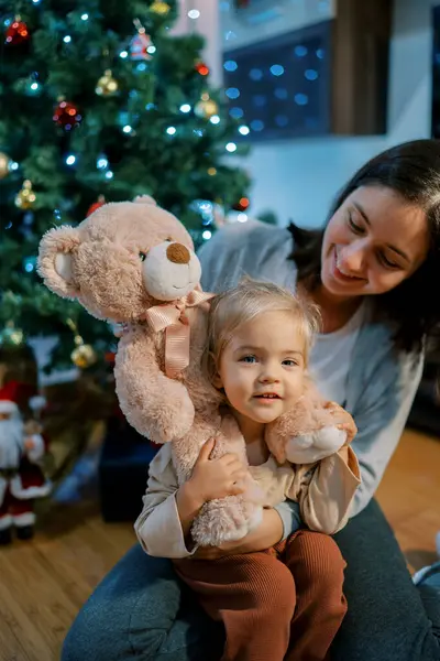 Mom put a teddy bear on the neck of a little girl sitting on the floor near the Christmas tree. High quality photo