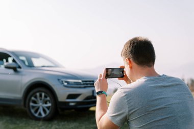 Sürücü, yeşil çimenlikte akıllı telefonuyla arabasının fotoğrafını çekiyor. Arkadan bak. Yüksek kalite fotoğraf
