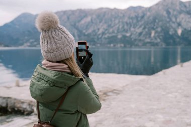 Kadın deniz kıyısında duruyor ve akıllı bir telefonla dağların fotoğraflarını çekiyor. Arkadan bak. Yüksek kalite fotoğraf