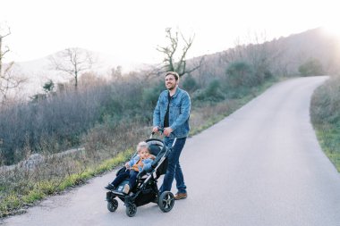 Bebek arabasında küçük bir kızla gülümseyen baba dağlarda yol boyunca yürüyor. Yüksek kalite fotoğraf