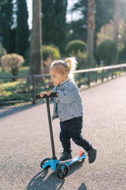 Küçük kız parkta bir yolda üç tekerlekli scooter 'a biniyor, ayağıyla itiyor. Yüksek kalite fotoğraf