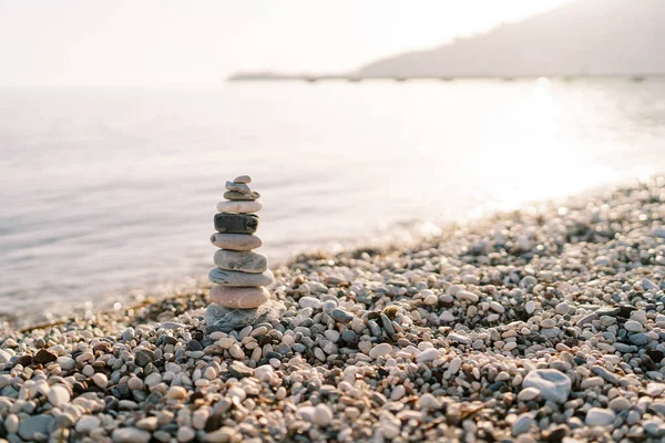 Cairn, parlak bir denizin yakınındaki çakıl taşlı bir sahilde, dağların arka planında duruyor. Yüksek kalite fotoğraf