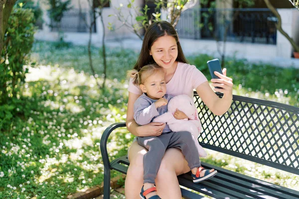 Gülümseyen anne, akıllı telefonuyla bankta kucağında düşünceli bir kızla selfie çeker. Yüksek kalite fotoğraf