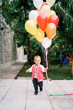 Bir sürü renkli balonu olan küçük bir kız yeşil bir bahçede yürüyor. Yüksek kalite fotoğraf