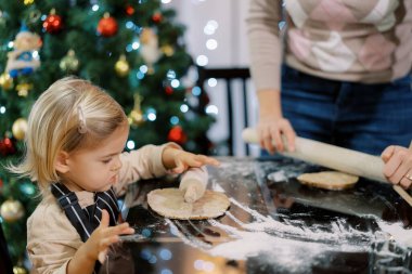 Küçük kız annesinin yanındaki masada merdaneyle hamuru yuvarlıyor. Yüksek kalite fotoğraf