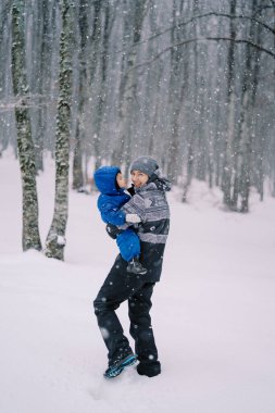 Kollarında küçük bir kızla gülümseyen anne bir kar yağışı altında ormanda kar yığınlarında yürür. Yüksek kalite fotoğraf