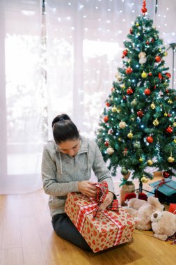 Genç bir kadın süslü bir Noel ağacının yanında oturur ve büyük bir hediye açar. Yüksek kalite fotoğraf