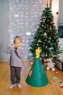 Küçük kız odada keçeli Noel ağacının yanında dururken kurabiye yiyor. Yüksek kalite fotoğraf