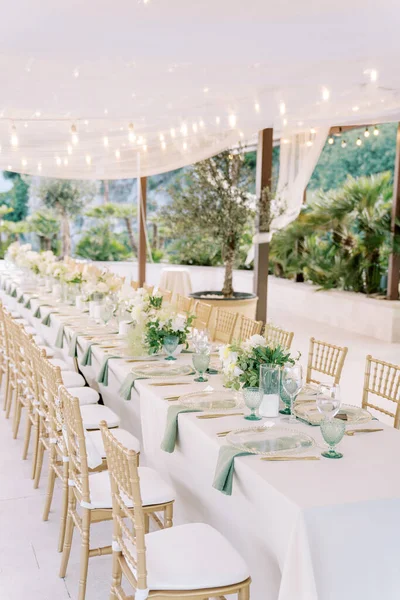 Long festive table with wicker chairs stands under a fabric canopy with lighting in the garden. High quality photo