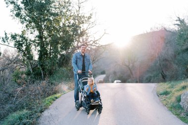 Gülümseyen baba, küçük kızını dağdan gelen yolda bebek arabasıyla taşıyor. Yüksek kalite fotoğraf