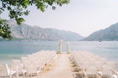 Beyaz sandalyeler, deniz kenarındaki bir rıhtımda, bir düğün kemerinin önünde, dağların arka planına karşı dururlar. Yüksek kalite fotoğraf