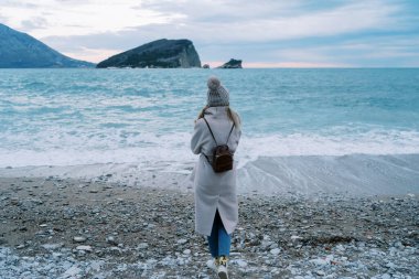 Sırt çantalı bir kız çakıl taşı plajında duruyor ve denize ve dağlara bakıyor. Arkadan bak. Yüksek kalite fotoğraf