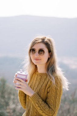 Güneş gözlüklü gülümseyen kız elinde bir fincan kahveyle bir dağda duruyor. Yüksek kalite fotoğraf