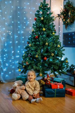 Küçük mutlu kız Noel ağacının yanında elinde oyuncak ayıyla yerde oturuyor. Yüksek kalite fotoğraf