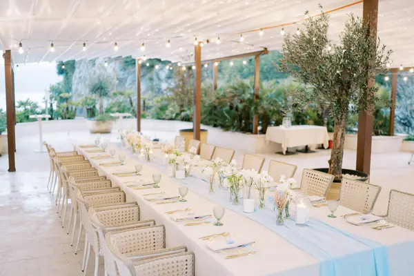 Festive long table with a strip of blue fabric in the middle under a canopy with glowing garlands. High quality photo