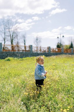 Karahindiba çiçekli bir çelenk içindeki küçük kız çiçek açan bir çimenlikte duruyor. Yan görüş. Yüksek kalite fotoğraf