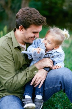 Küçük kız yeşil çimlerde babasının kucağında elinde oyuncakla oturuyor. Yüksek kalite fotoğraf
