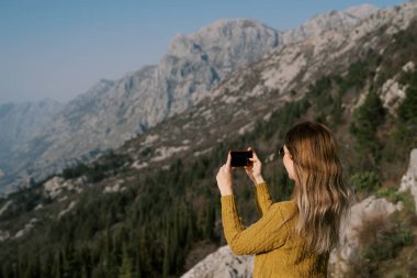 Genç bir kadın bir dağda duruyor ve akıllı telefonuyla manzara çekiyor. Yan görüş. Yüksek kalite fotoğraf