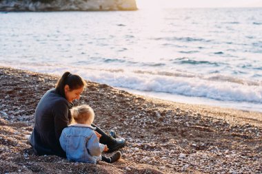 Anne ve küçük kız sahilde oturuyor ve çakıl taşlarıyla oynuyorlar. Arkadan bak. Yüksek kalite fotoğraf