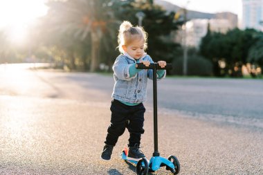 Küçük kız scooter 'a biniyor, ayağını yola koyuyor, tekerleklerin altına bakıyor. Yüksek kalite fotoğraf