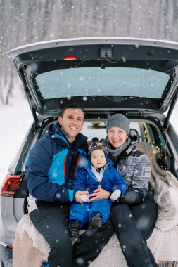 Gülümseyen anne, baba ve küçük kız kış ormanlarında arabanın bagajında battaniyenin üzerinde oturuyorlar. Yüksek kalite fotoğraf