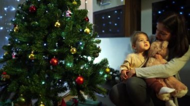 Annem kollarında küçük bir kızla odadaki süslü bir Noel ağacının yanında oturuyor. Yüksek kalite 4k görüntü