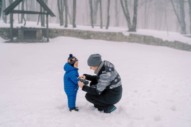 Annem karlı bir parkta çömelmiş küçük bir çocuğa eldiven takıyor. Yüksek kalite fotoğraf