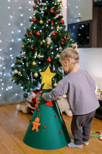 Küçük kız oyuncak asıyor, keçeli Noel ağacının yanında duruyor. Arkadan bak. Yüksek kalite fotoğraf