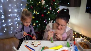 Anne ve küçük kızı Noel ağacının yanındaki masada kurabiye süslüyor. Yüksek kalite 4k görüntü