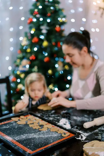 Tray of cut-out cookies stands on a table next to a mother and little daughter making cut-outs. High quality photo