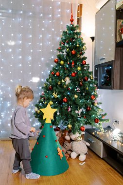 Küçük kız keçeli Noel ağacını oyuncaklarla süslüyor, odada duruyor. Yüksek kalite fotoğraf