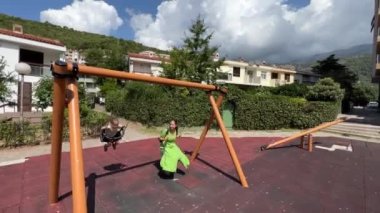 Annem ve küçük bir kız evlerin yanındaki parkta salıncakta sallanıyorlar. Yüksek kalite 4k görüntü