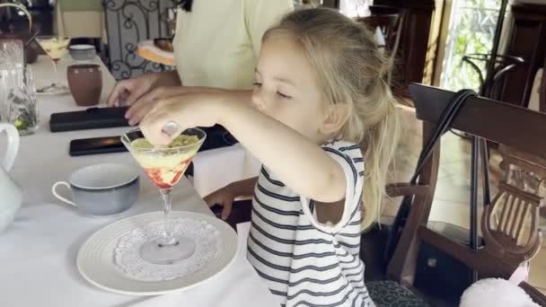 小さな女の子はガラスのスプーンでレストランのテーブルでアイスクリームを食べます 高品質4K映像 — ストック動画