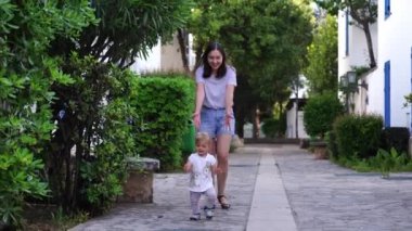 Gülümseyen anne, avluda asfalt yolda ilk adımlarını atan küçük bir kızı takip ediyor. Yüksek kalite 4k görüntü