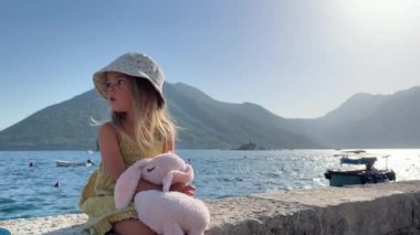 Küçük kız denizin kenarındaki taştan bir çitte oturuyor. Dağların arkasına bakıyor. Yüksek kalite 4k görüntü