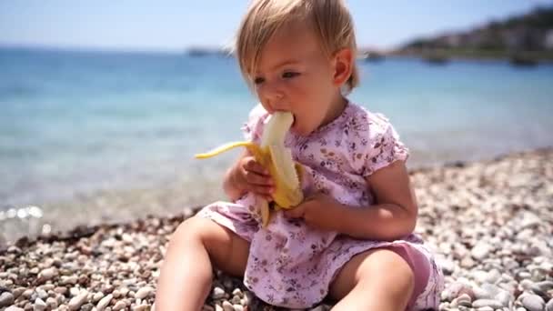 Kislány Banánt Eszik Miközben Kavicsos Tengerparton Üldögél Kiváló Minőségű Felvételek — Stock videók